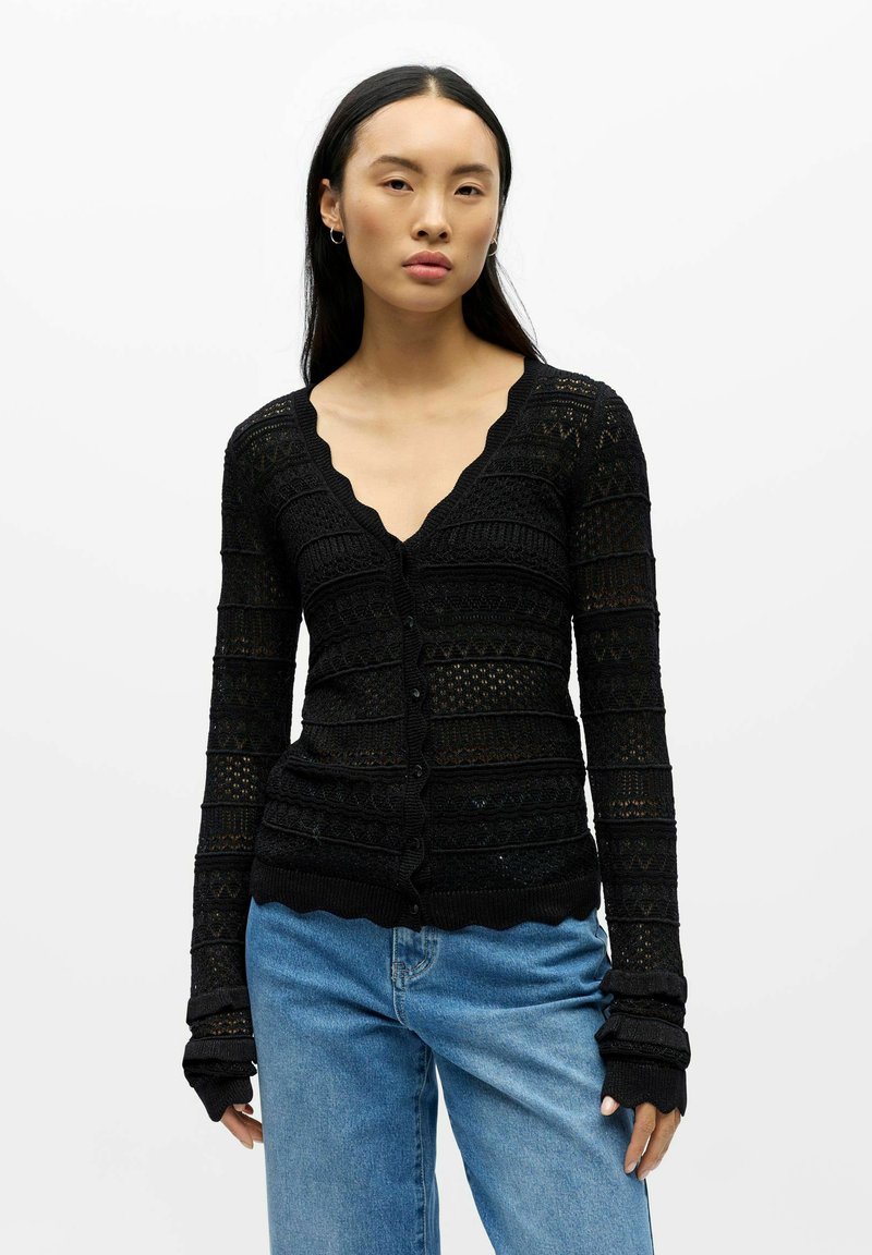 Schwarze Strickjacke mit V-Ausschnitt, strukturiertem Strickmuster und gewellten Kanten. Hat lange Ärmel und einen Knopfverschluss. Kombiniert mit hellblauen Jeans.