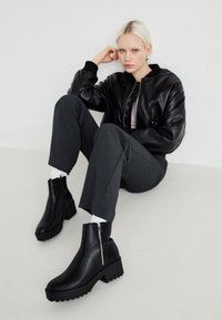 Botas de cuero negras hasta el tobillo con un tacón grueso, que cuentan con un detalle de cremallera en el costado y una suela texturizada, combinadas con pantalones grises y una chaqueta bomber negra.