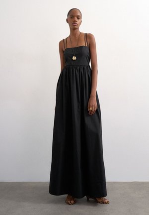 Femme portant une robe maxi noire sans manches à fines bretelles, un collier en or, des bagues en or et des sandales à talons hauts à imprimé animal, debout à l'intérieur.