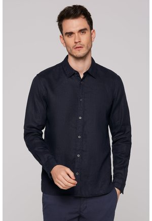 Mann mit kurzen dunklen Haaren, der ein geknöpftes, dunkelblaues Hemd mit langen Ärmeln trägt und vor einem schlichten hellen Hintergrund steht.