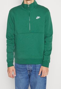 Green Nike sweatshirt featuring a half-zip collar, soft fabric, ribbed cuffs, and a white logo on the left chest. Paired with blue jeans.