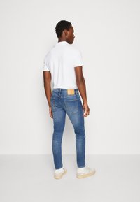 Slim fit, világoskék farmernadrág hátsó zsebekkel és bézs bőr logóval. Fehér sportcipőkkel párosítva.