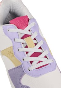 Zapatilla blanca con acentos morados, rosas y amarillos. Destacan los materiales texturizados, el cordón prominente y el logo de 'GAP' en la lengüeta.
