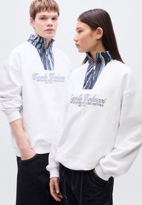 Λευκό φούτερ με ψηλό γιακά και επένδυση με μπλε και λευκό μοτίβο. Κεντημένο λογότυπο μπροστά. Το μοντέλο ποζάρει φορώντας σκούρο τζιν.