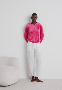 Fato de moletom rosa de veludo com fecho éclair e bolsos frontais, combinado com calças largas brancas e texturizadas. Com fundo neutro.
