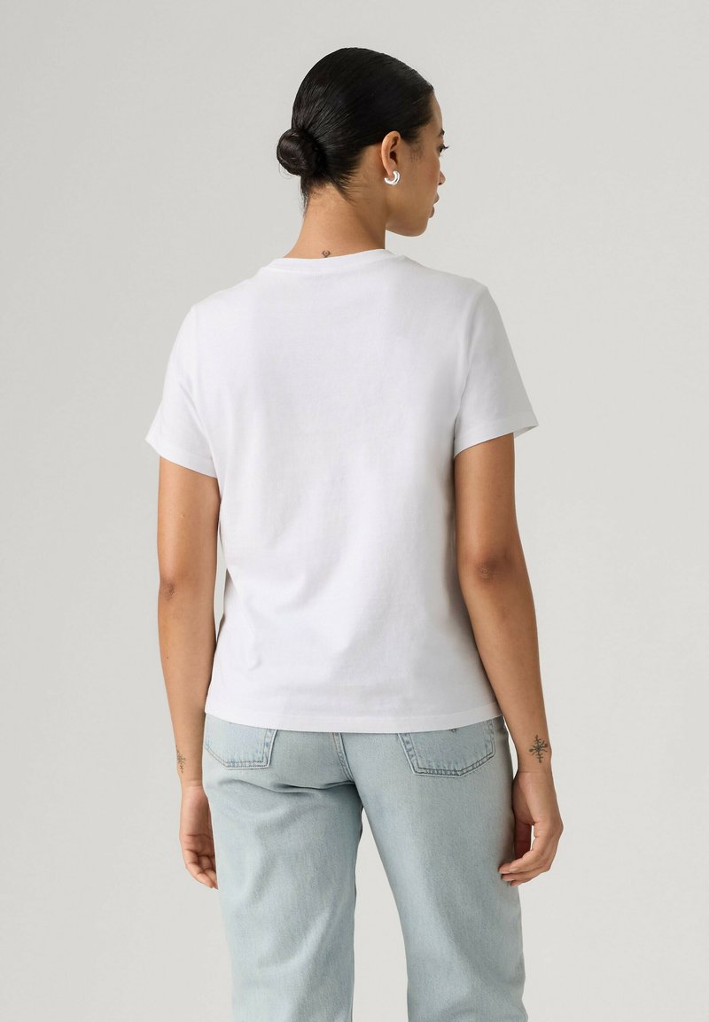 Femme aux cheveux foncés en chignon, portant des boucles d’oreilles anneaux argentés, un t-shirt blanc uni à manches courtes et un jean bleu clair, vue de dos.
