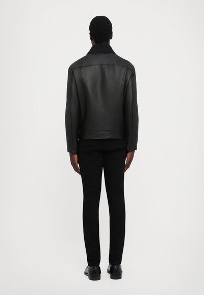Man staat met zijn rug naar de camera en draagt een zwarte leren jas, zwarte pantalon en zwarte nette schoenen tegen een simpele witte achtergrond.