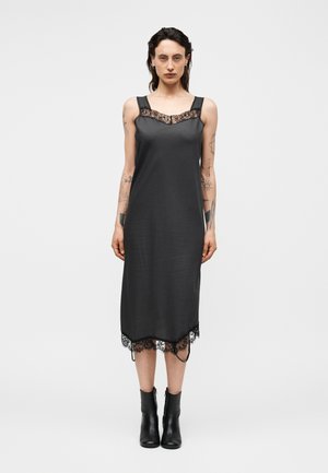 Donna con tatuaggi che indossa un vestito midi nero senza maniche con rifiniture in pizzo e stivaletti neri, in piedi su uno sfondo bianco semplice.