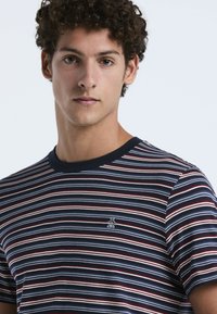 Jonge man met krullend haar, gekleed in een marineblauw, wit en rood gestreept T-shirt met een klein pinguïnlogo op de borst, die recht vooruit kijkt.