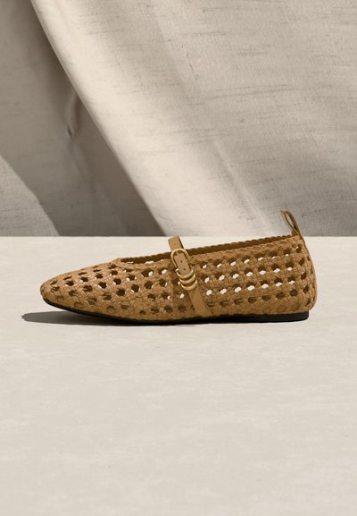 Καφέ ανοιχτό παπούτσι από πλεκτό ύφασμα με λεπτό λουράκι με αγκράφα και μικρό κρίκο στη φτέρνα, εκτεθειμένο σε φωτεινή επιφάνεια με μπεζ υφασμάτινο φόντο.