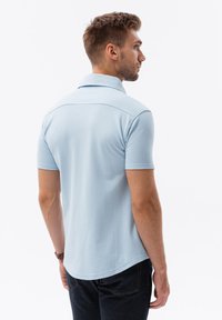 Camisa polo azul claro con mangas cortas, cuello y dobladillo curvo. Textura suave con un diseño sencillo, que presenta un detalle en la yugo trasero.