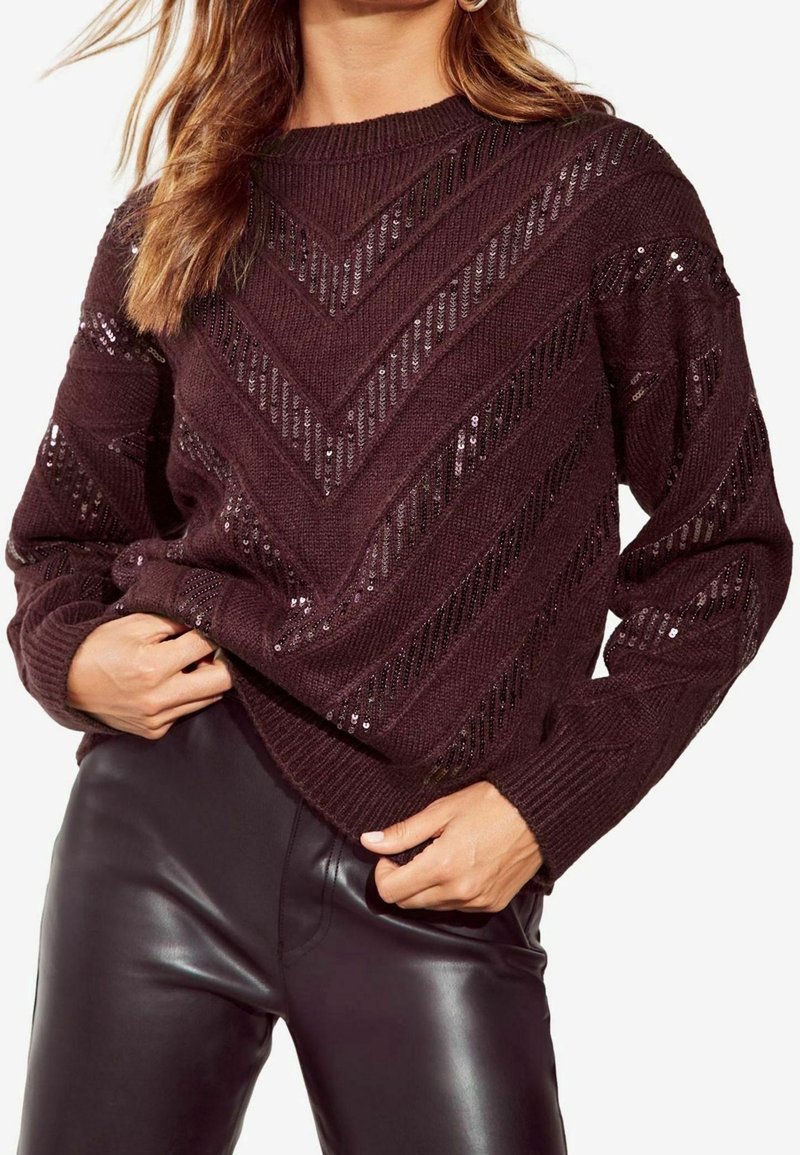 Femme portant un pull en maille marron foncé avec des motifs chevrons en sequins et un pantalon noir en simili cuir, mains ajustant l'ourlet du pull.