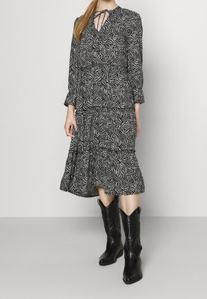Schwarzes und weißes gemustertes Kleid mit langen Ärmeln, Schleifenkragen, rüschengesäumtem Saum, kombiniert mit schwarzen Cowboy-Stiefeln. Glatter Stoff und floralem Design.