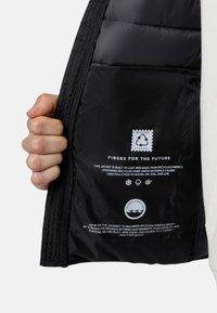 Gepolsterde zwarte jackinterieur met een satijnachtige textuur, met een label voor gerecyclede materialen en milieuboodschappen. Een hand grijpt de rits.