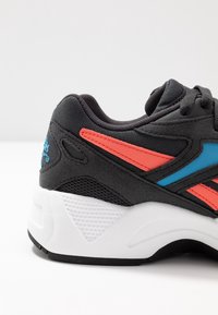 Czarny but sportowy z szarymi i pomarańczowymi akcentami, siateczkowymi panelami i białą, teksturowaną podeszwą. Posiada zaokrąglony czubek i system sznurowania.