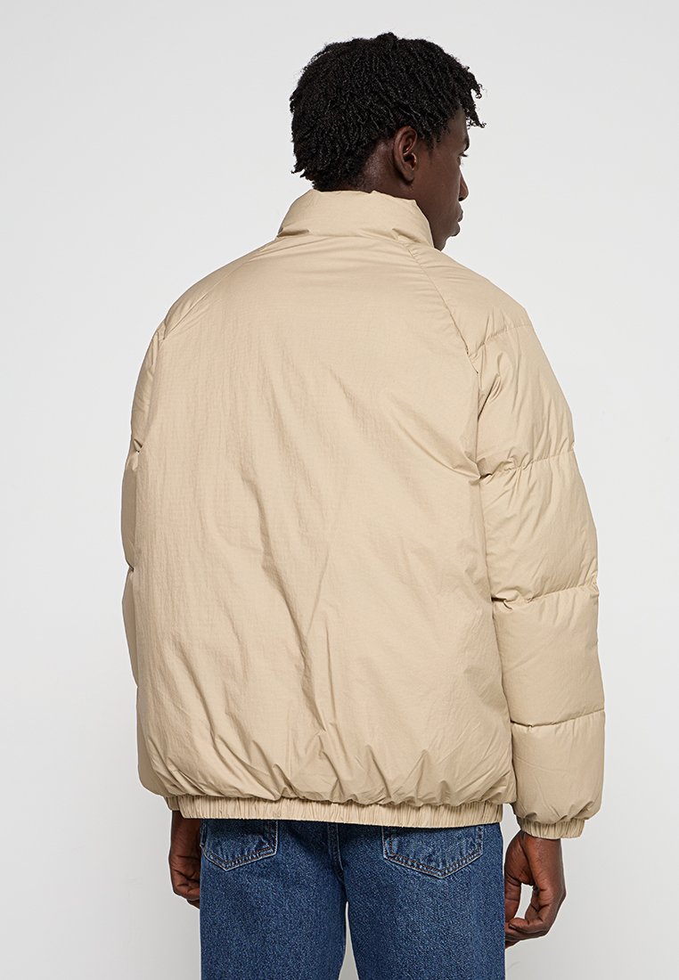 Chaqueta de plumas beige con un cuello alto, con textura acolchada, dobladillo elástico y costuras visibles en la parte posterior. Lleva puesta sobre unos jeans azules.
