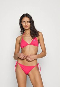 Sieviete ar gariem tumšiem matiem, tērpusies spilgti rozā aukliņu bikini ar sānu saitēm, stāv uz viendabīga baltā fona.