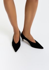 Černé semišové slingback vysoké podpatky s špičatou špičkou, mají nízký podpatek a hluboký V-výstřih v přední části. Hladká textura.