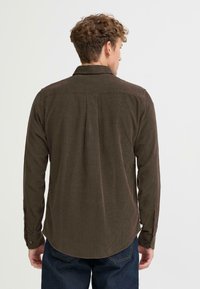 Camisa de pana marrón con mangas largas, puños abotonados y una hemline redondeada; presenta una textura de canalé vertical y un detalle de pliegue en la parte posterior.