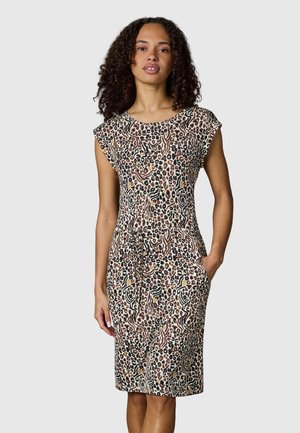 Nainen seisoo pukeutuneena hihattomaan polvipituiseen mekkoon, jossa on monivärinen leopardikuosi ja abstrakti eläinkuosi, toinen käsi taskussa.