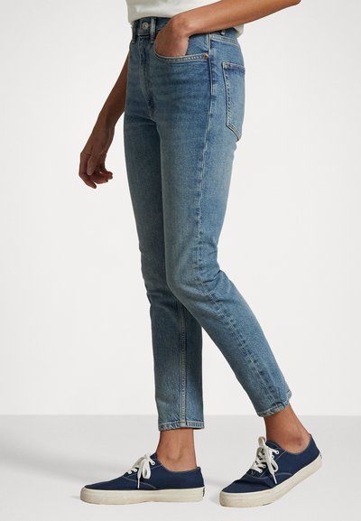 Pessoa vestindo jeans slim-fit azul claro e ténis de lona azul marinho com atacadores brancos, em pé com uma mão no bolso.