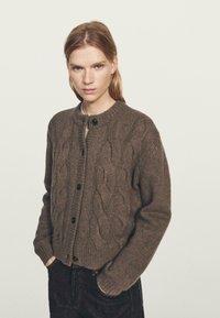 Bruine gebreide cardigan met kabelbreipatroon, ronde halslijn en knoopsluiting, voorzien van lange mouwen en een zachte, gestructureerde afwerking.