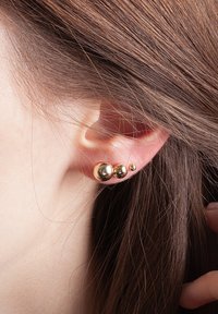 Złote kolczyki z trzema sferycznymi elementami o różnych rozmiarach, o gładkiej, odbijającej powierzchni, noszone w przekutym uchu.