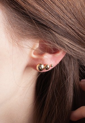 Złote kolczyki z trzema sferycznymi elementami o różnych rozmiarach, o gładkiej, odbijającej powierzchni, noszone w przekutym uchu.