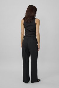 Frau steht mit dem Rücken zur Kamera, trägt ein schwarzes ärmelloses Oberteil, hochtaillierte, weit geschnittene schwarze Hosen und spitze schwarze Schuhe vor einem grauen Hintergrund.