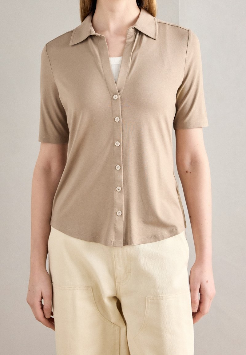 Chemise courte à manches courtes de couleur brun clair avec col et fermeture à boutons, en tissu doux, portée avec un pantalon taille haute de couleur crème.