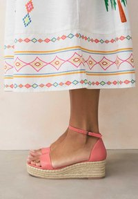 Sandales compensées en cuir rose avec une plateforme en corde de jute, dotées d'une bride de cheville et d'un large bandeau à l'avant. Jupette blanche à ourlet brodé en accompagnement.