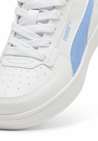 Baltos odinės sportinės bateliai su šviesiai mėlynais akcentais, apvaliais perforacijomis ir plokščiomis baltomis raištelėmis. Šone yra šviesiai mėlyna Puma logotipas.