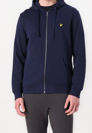 Mann trägt einen marineblauen Kapuzenpullover mit Reißverschluss, Fronttaschen und einem kleinen gelben Vogel-Logo auf der linken Brust.
