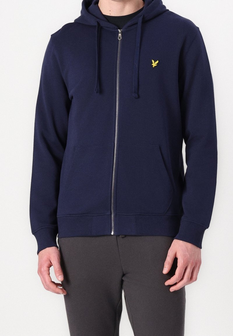 Homme portant un sweat à capuche bleu marine zippé avec des poches avant et un petit logo jaune d'oiseau sur la poitrine gauche.