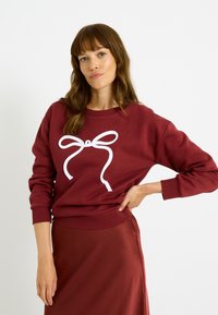 Sudadera color burdeos con un gran gráfico de lazo blanco en la parte frontal. El material parece suave con puños y dobladillo acanalados.