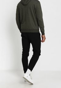 Man som bär en mörkgrön hoodie, svarta byxor, vita strumpor och vita sneakers, står mot en enkel ljus bakgrund och vänder sig bort.