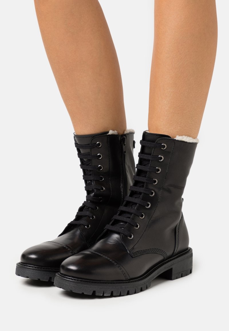 Bottines noires en cuir à lacets avec semelles épaisses et doublure en polaire blanche, portées sur des jambes nues sur fond blanc.