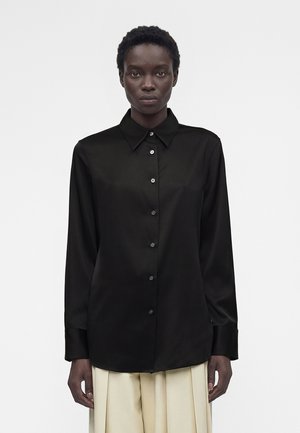 Personne portant une chemise noire à manches longues avec boutons et col pointu, et un pantalon plissé beige clair, debout devant un fond uni.