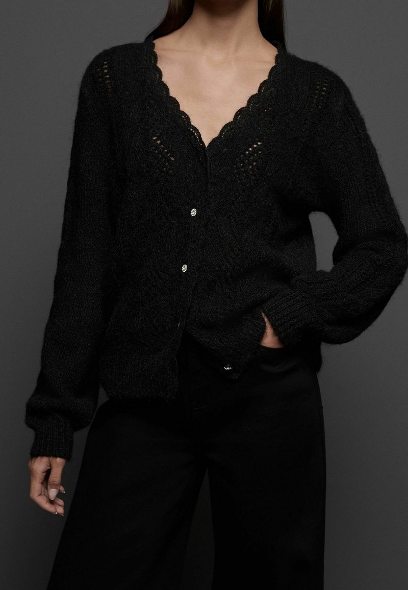 Femme portant un cardigan noir en maille avec des bords festonnés et des boutons brillants, associé à un pantalon noir, sur un fond sombre.
