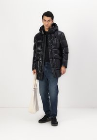 Geacă pufă neagră cu glugă, textură lucioasă, design matlasat, închisă cu nasturi, asortată cu blugi albaștri și pantofi negri, ținând o geantă tip tote.