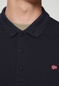 Polo bleu marine avec une finition texturée, comprenant une patte de boutonnage à trois boutons et un logo drapeau rouge et blanc brodé sur la poitrine.