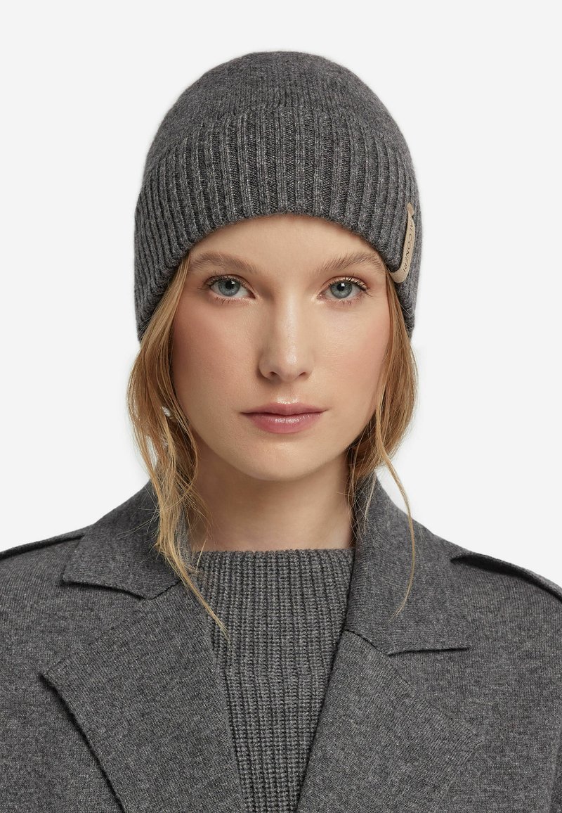 Graue Strickmütze mit gerippter Textur und einem dezenten Logo-Patch. Das Material wirkt weich und warm, geeignet für kaltes Wetter.