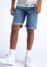 Blauwe denimshorts met omgeslagen zoom, vijf-pocketstijl en gele stikdetails. Gecombineerd met witte sneakers en witte crew-sokken.