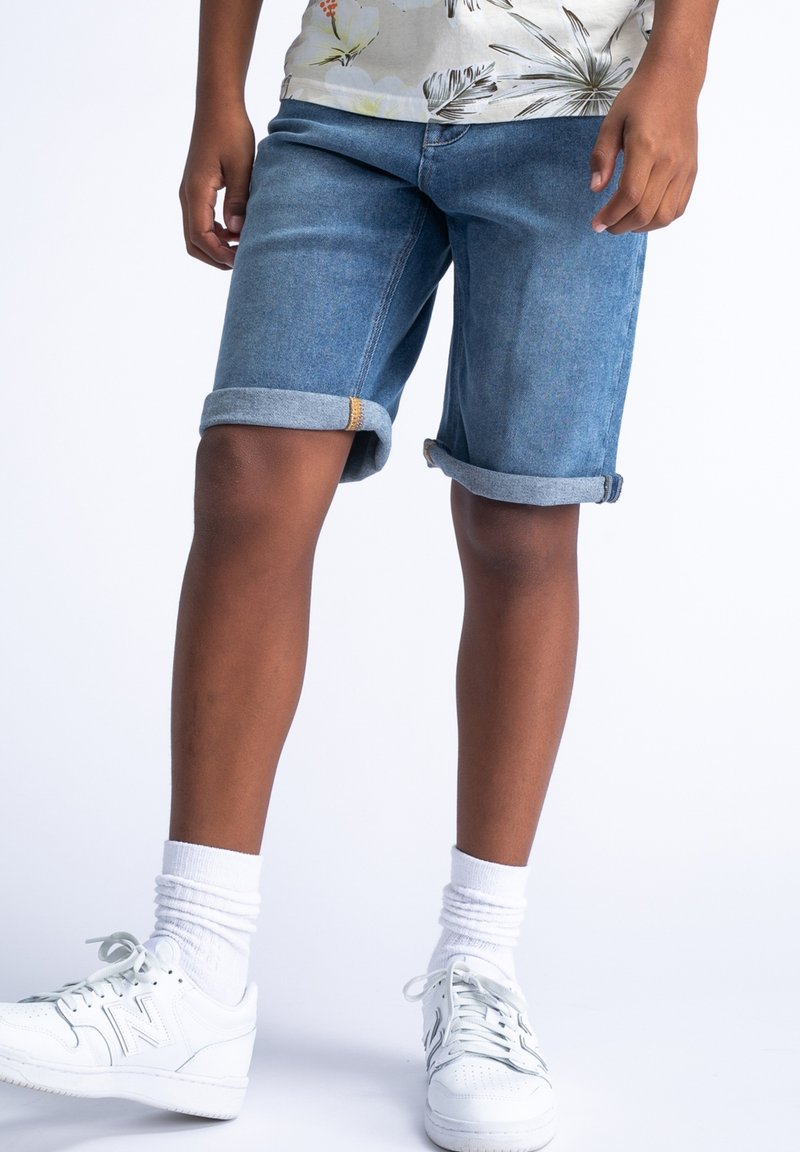 Blauwe denimshorts met omgeslagen zoom, vijf-pocketstijl en gele stikdetails. Gecombineerd met witte sneakers en witte crew-sokken.