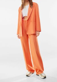 Oranžový blazer a odpovídající široké kalhoty, vyrobené z hladkého materiálu, s dvojitým zapínáním na dva knoflíky a bočními kapsami.