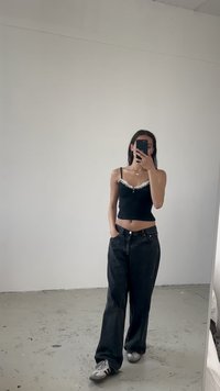 Čierny top bez rukávov s čipkovým lemovaním a detailom gombíkov, skombinovaný s voľnými čiernymi džínami. Sivé tenisky s viditeľnými vzorovanými akcentmi. Reflexia v zrkadle na celú postavu.