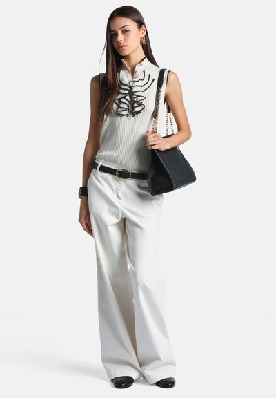 Jeune femme portant un haut blanc sans manches avec un motif brodé noir, un pantalon large blanc, une ceinture noire, des chaussures noires, et tenant un sac à main noir.