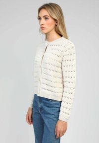 Cardigan en tricot ivoire avec un design ajouré, un col rond et une fermeture à boutons. Il présente des manches longues et un ourlet court. Tissu texturé.