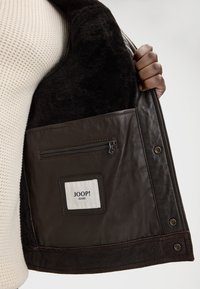 Wnętrze brązowej skórzanej kurtki z miękką czarną podszewką, jedną kieszenią na zamek oraz metką z napisem "JOOP! JEANS".