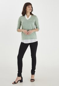 Lys grøn, struktureret sweater med v-hals og trekvartærmer over en hvid lagdelt skjorte, parret med sorte bukser og sorte hæl-sandaler.